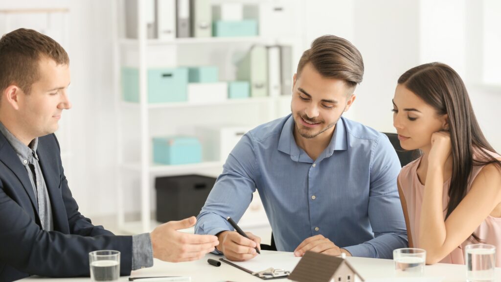 Young couple sitting with a real estate agent, reviewing and signing property sale documents in an office to comply with QLD Form 2 seller disclosure.
