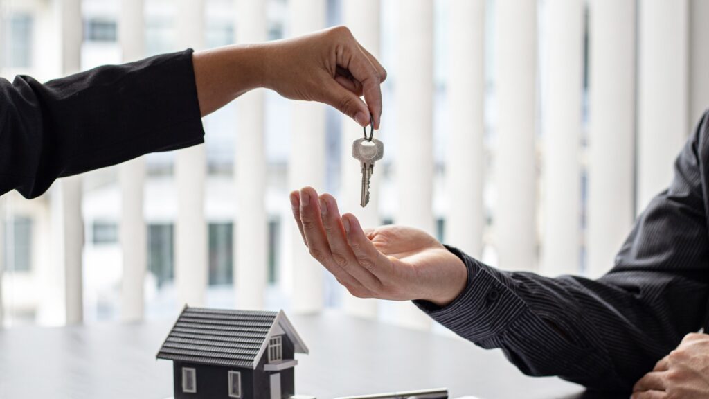 A person handing over a set of house keys to another person, with a small model home on the table, symbolising a rental or property management agreement.