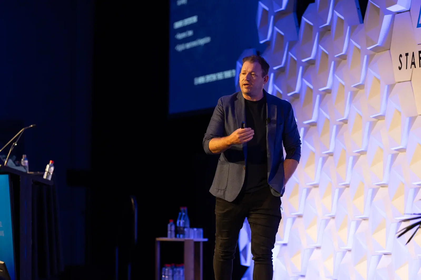 Conference speaker presenting on stage at the STAFFLINK Conference with a presentation screen and branded geometric backdrop.